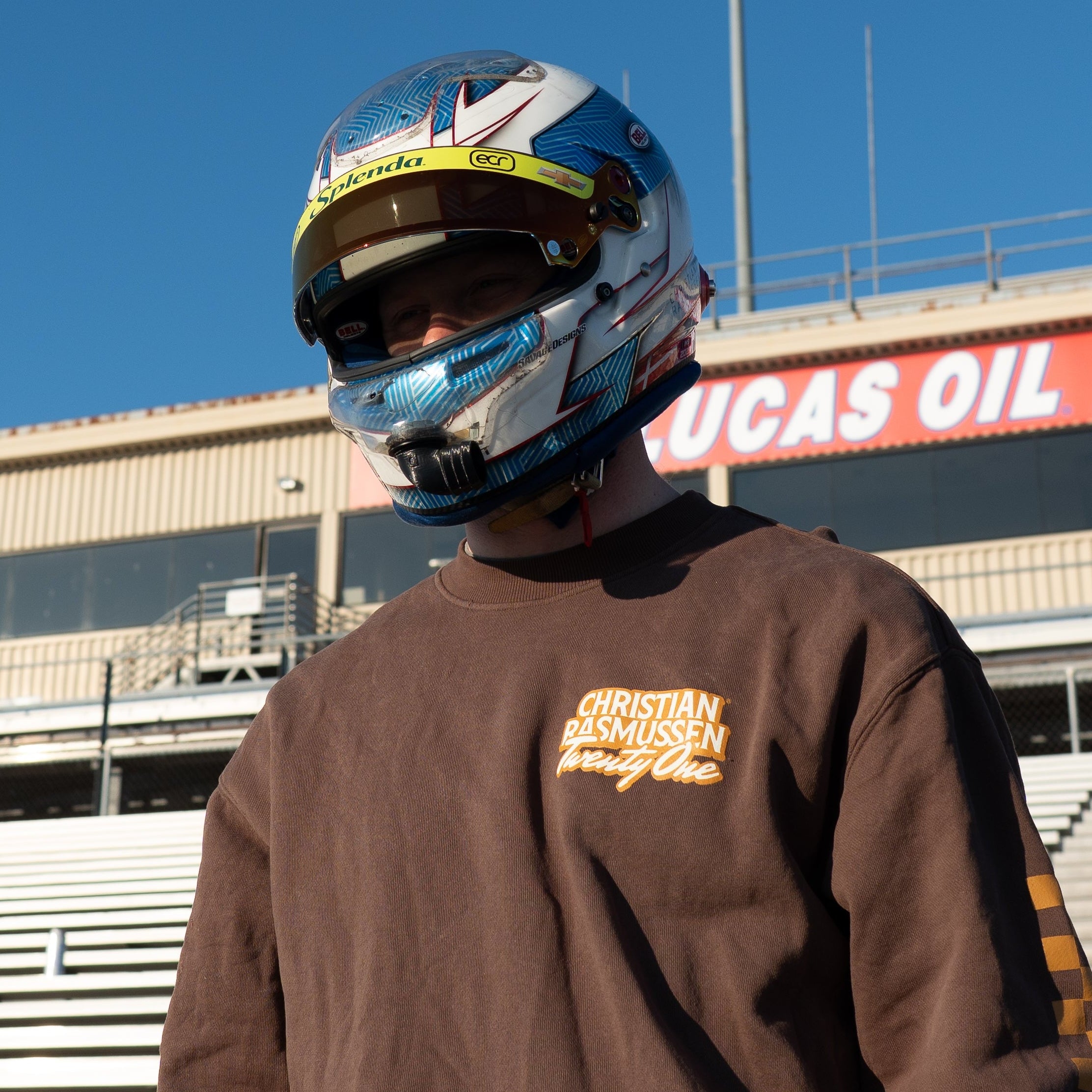 Person wearing a racing helmet and brown sweatshirt with a Lucas Oil sign in the background