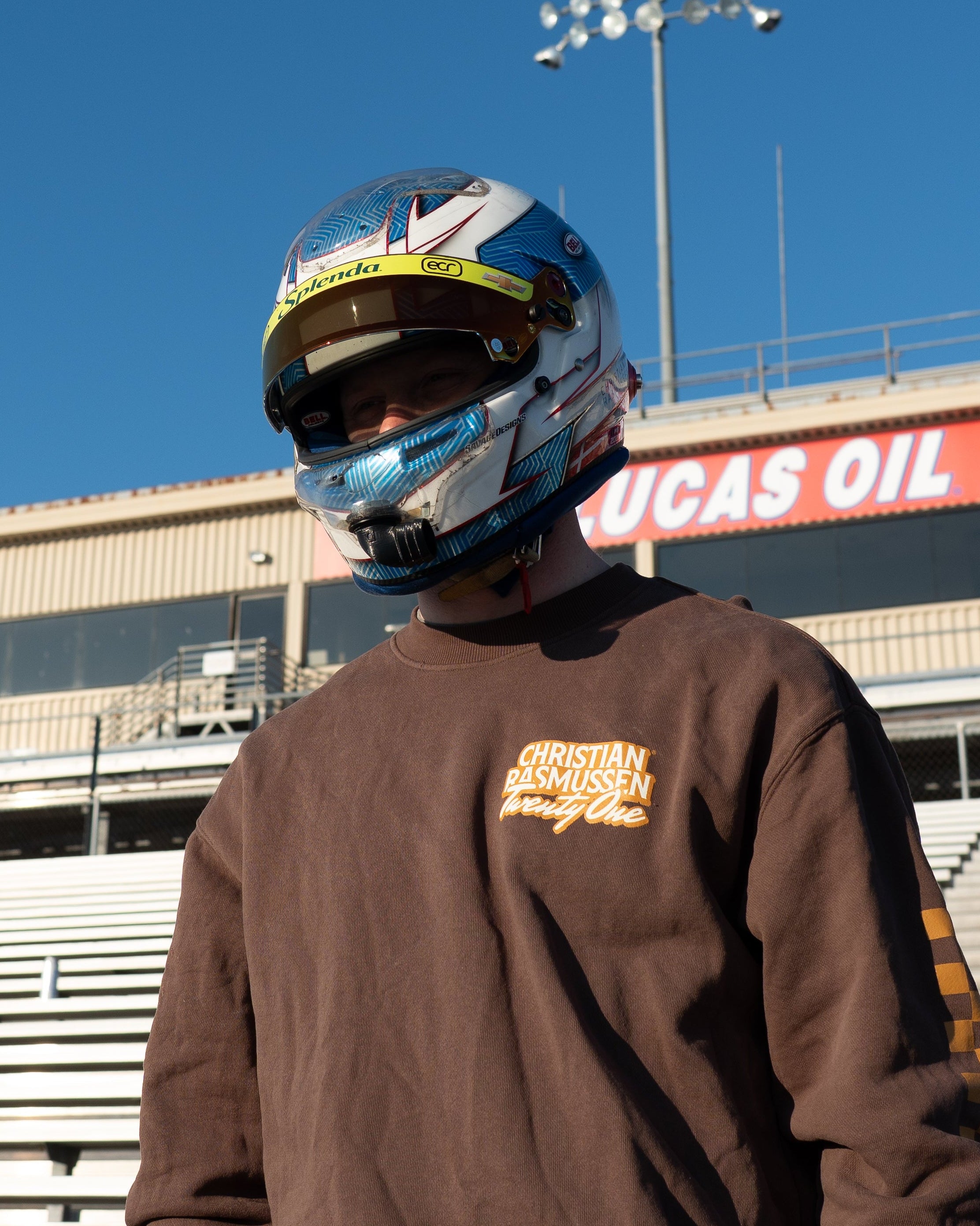 Person wearing a racing helmet and brown sweatshirt with a Lucas Oil sign in the background