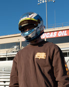 Person wearing a racing helmet and brown sweatshirt with a Lucas Oil sign in the background
