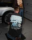 Two people working on a car in a garage, with one person wearing a branded t-shirt.