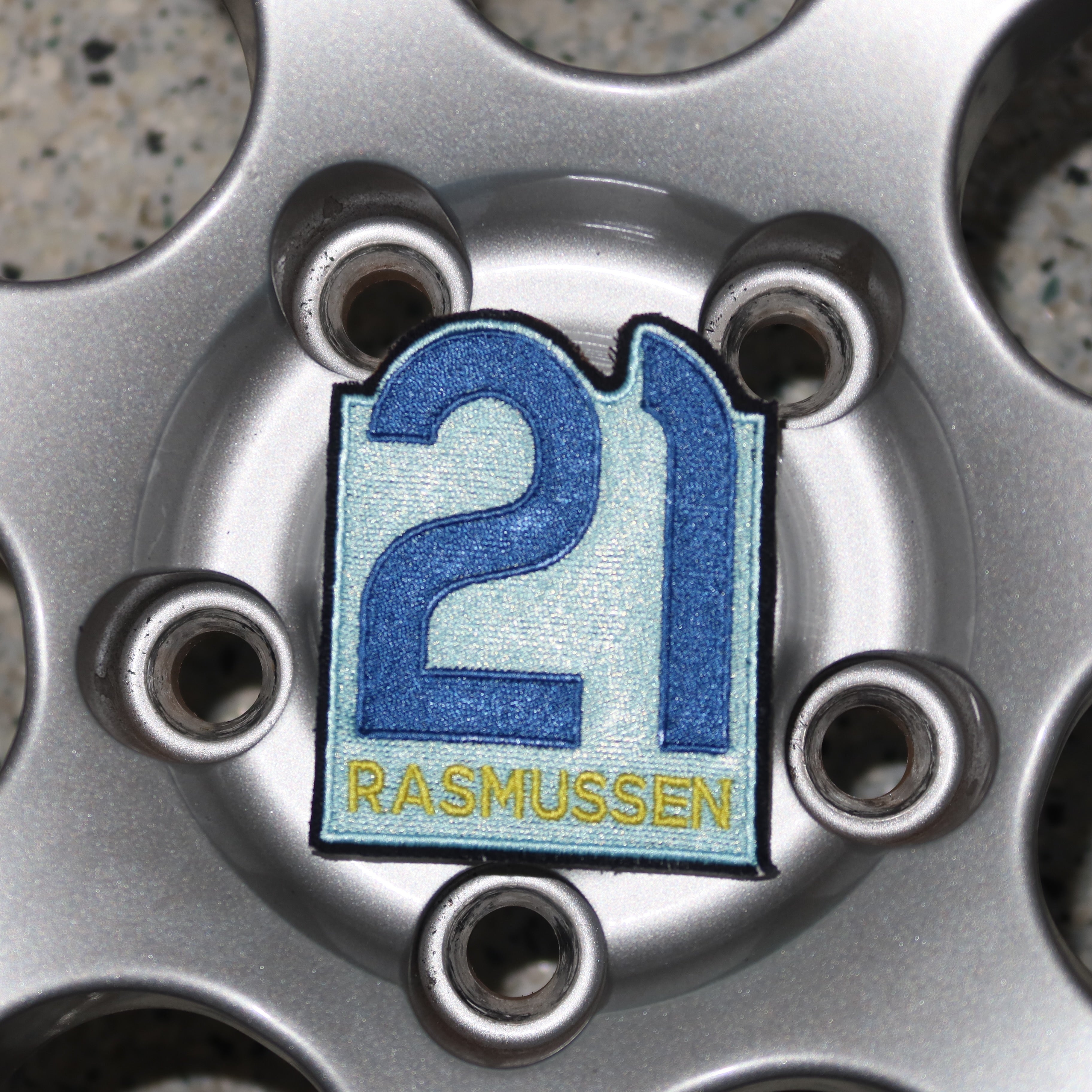 Close-up of a car wheel with a '21 Rasmussen' badge on a textured pavement background
